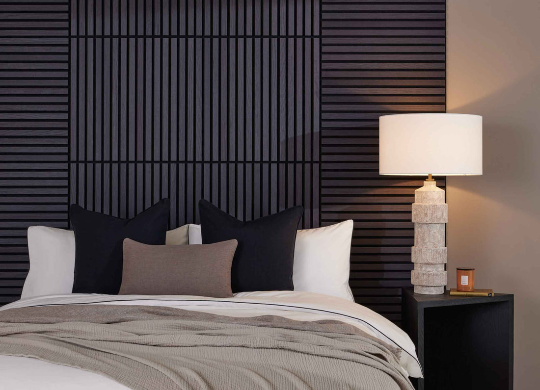 Bedroom with black wall panelling, a white bed, black and brown cushions, a beige throw, black side table and a stone-effect lamp.