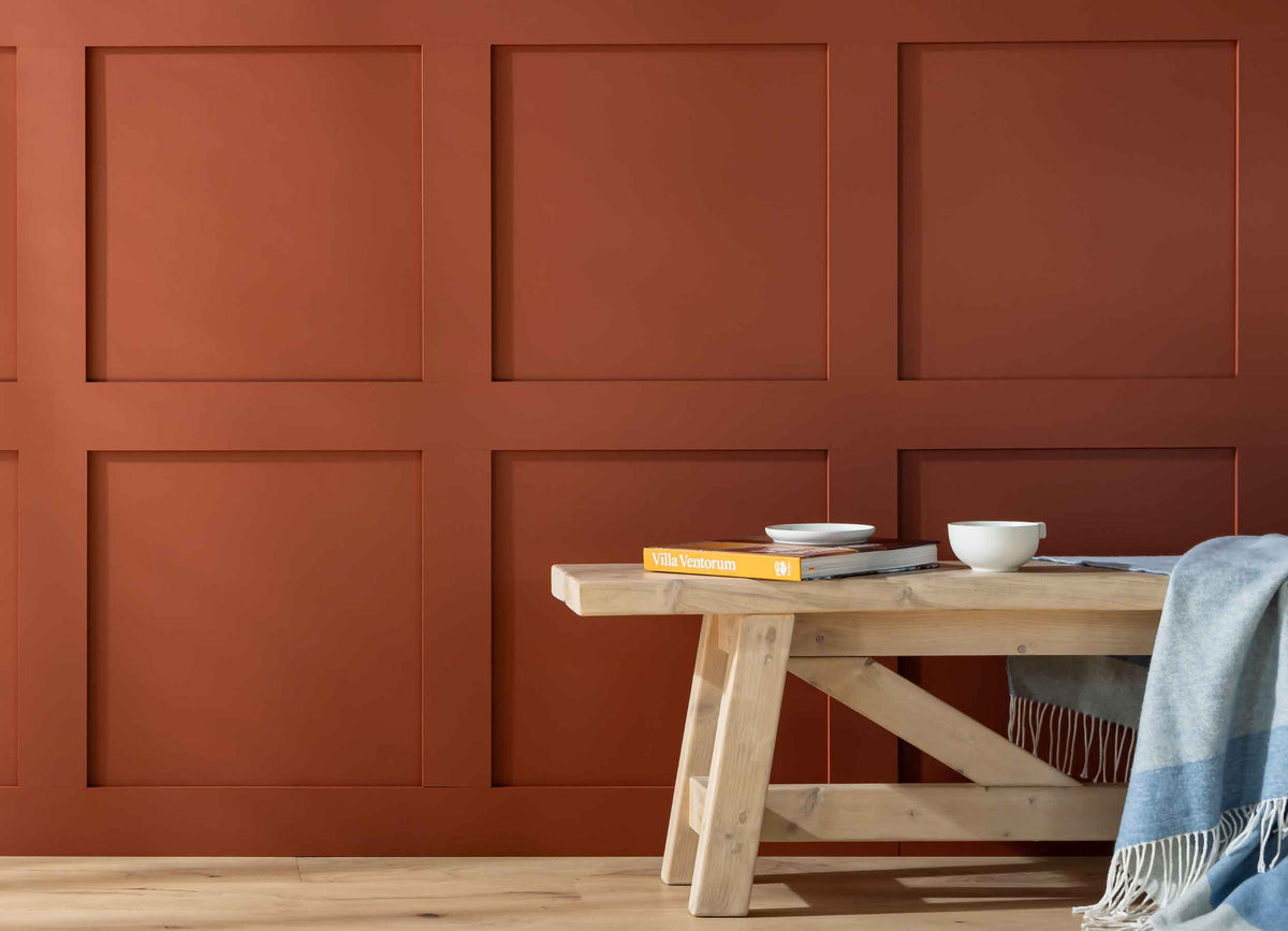 Terracotta square Shaker-style wall panelling behind a pine bench with a blue throw, an orange book and a white cup and saucer.