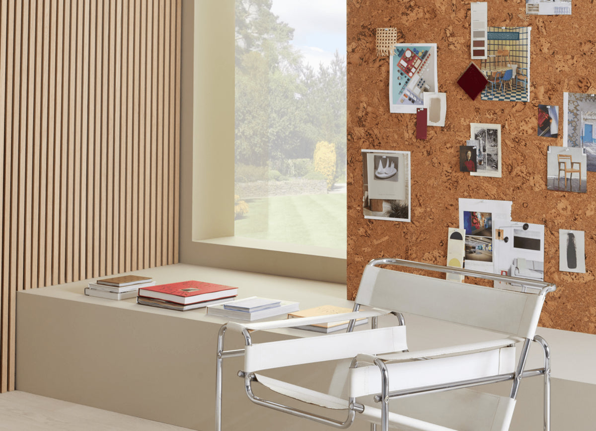 A cork pinboard featuring pictures behind a row of books and a white and silver chair, next to a window and SlatWall panels.