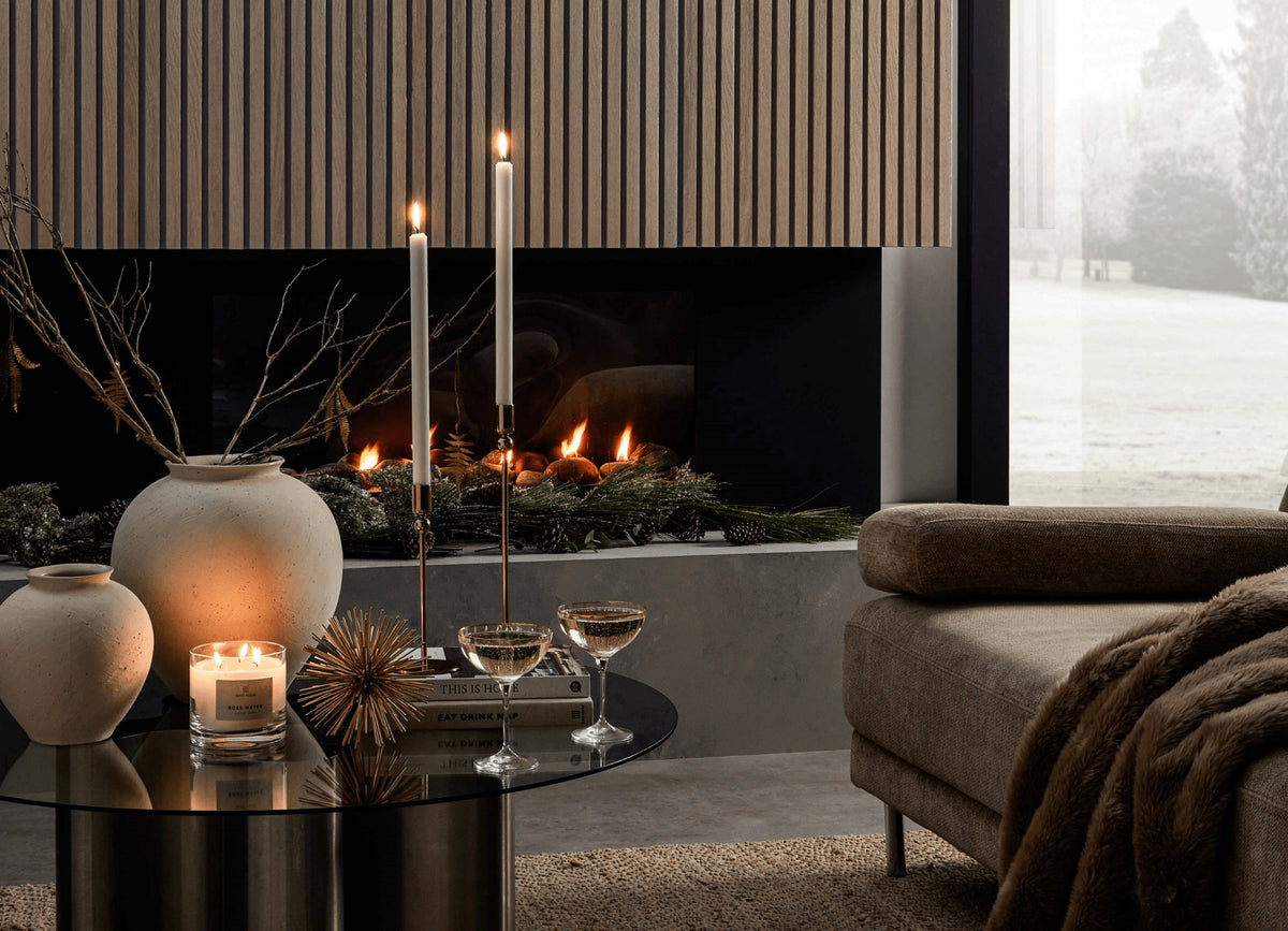 This image shows a fireplace with a SlatWall covering, in front of the fire there is a coffee table with lit candles, two glasses or champagne and some Christmas ornaments. The corner of a sofa can be seen also.