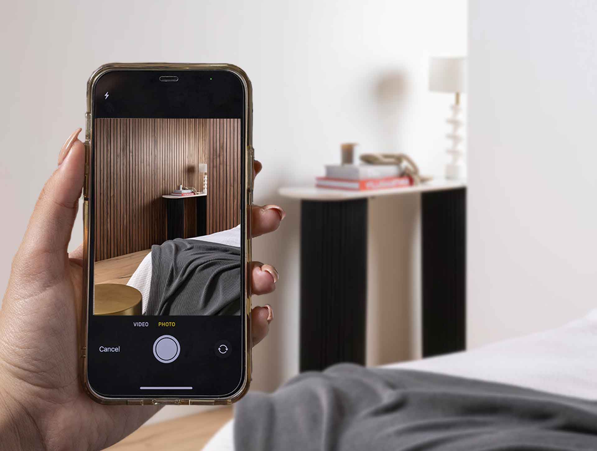 Hand holding phone with image of deep walnut SlatWall on wall with bed with grey and white sheet. Sideboard with books and lamp. Image in backgroung is the same as phone with no SlatWall on the walls, instead blank white